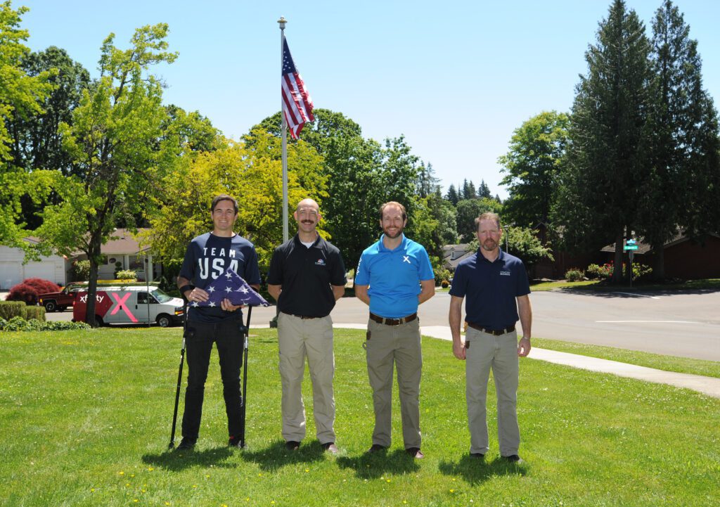 People stand in a field with an American flag on top of a flag pole in the background.